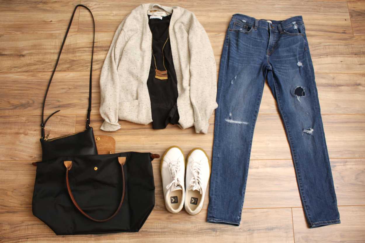 A pair of jeans, a tank, a cardigan, sneakers, and two purses are arranged on the floor in a flatlay