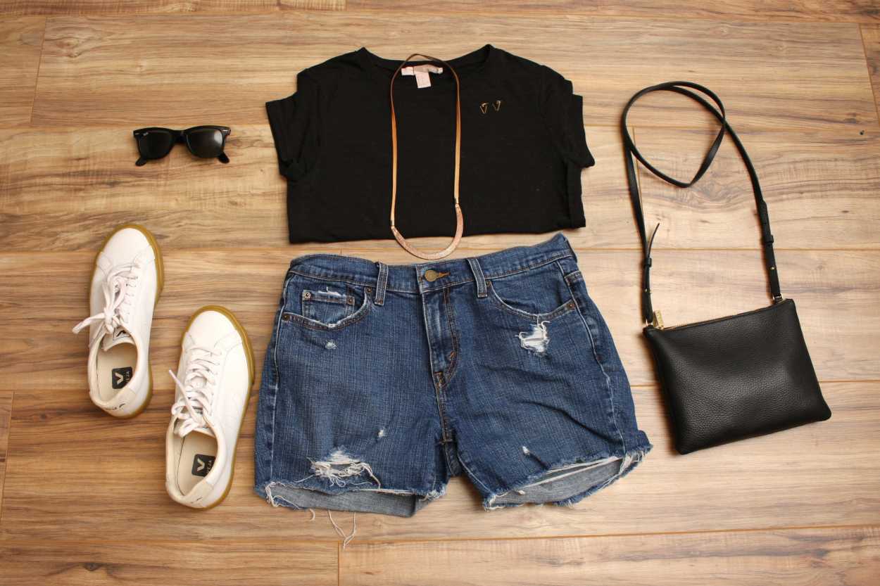 A tee, shorts, sneakers, and bag are arranged on a wood floor in a flatlay