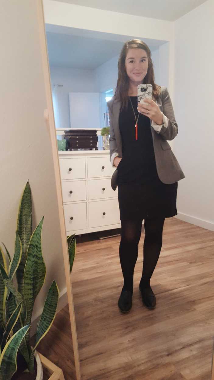 Alyssa wears a black dress, black tights, black shoes, and grey blazer and smiles at the mirror