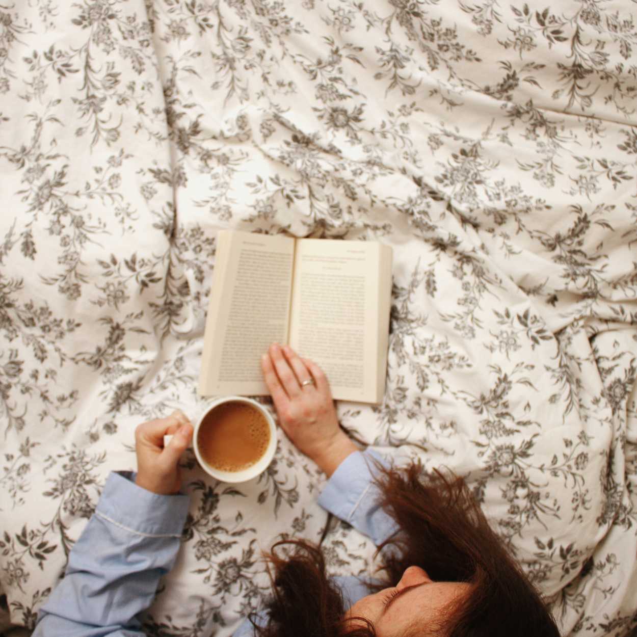 Alyssa sits on a bed reading a book with a cup of coffee