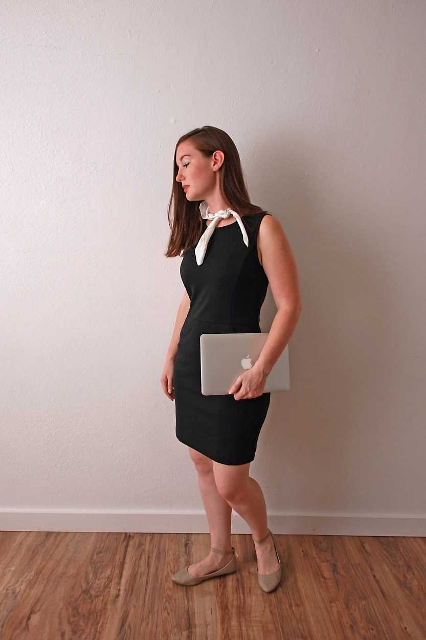Alyssa wears a black shift dress, beige flats, and a neck scarf and holds a laptop