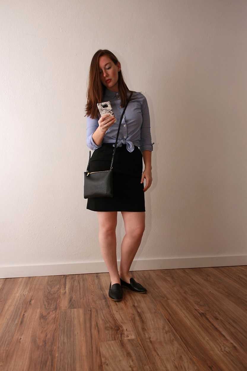 Alyssa wears a blue button-down tied in a knot over a black dress with loafers and looks at her phone
