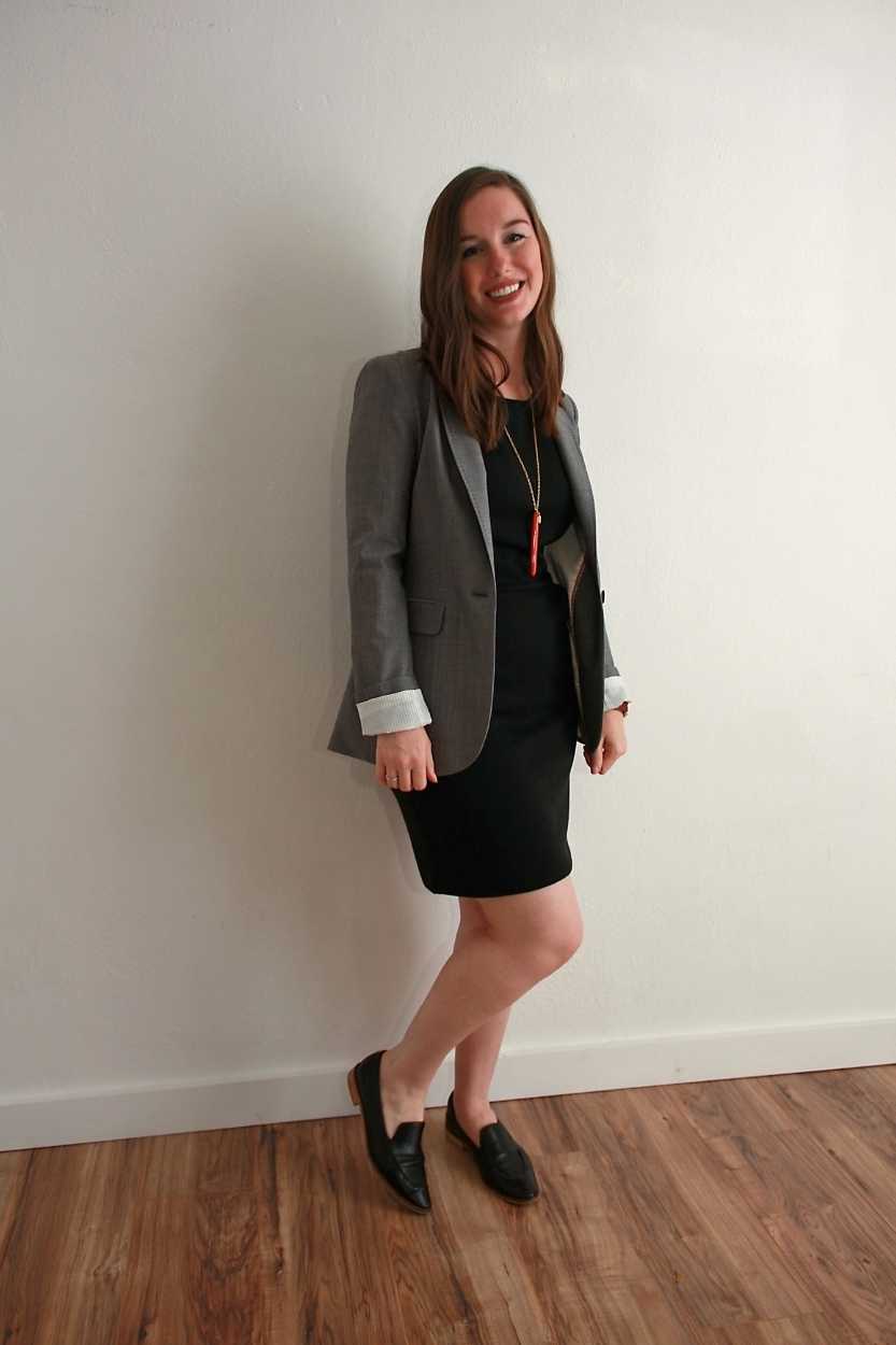 Alyssa wears a black dress, black shoes, and grey blazer and smiles at the camera