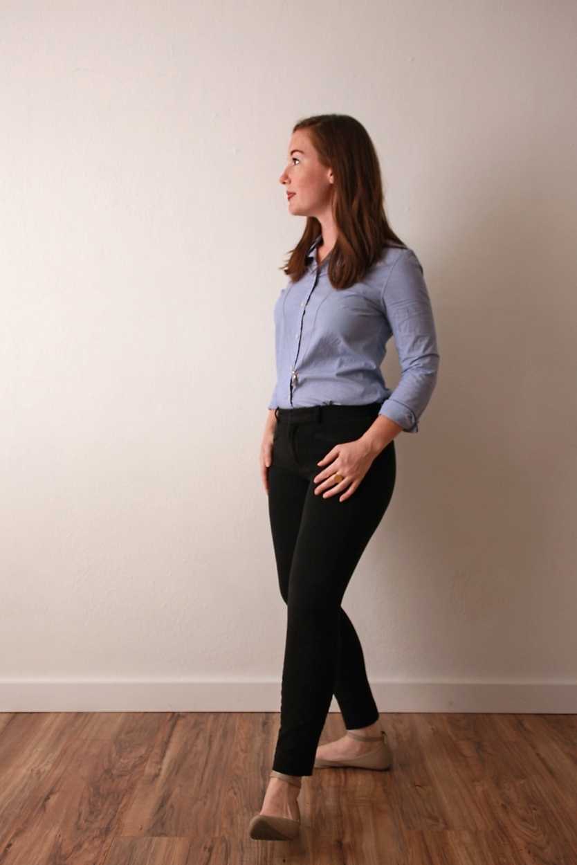 Alyssa wears a blue oxford shirt with black pants and beige flats and faces sideways to the camera