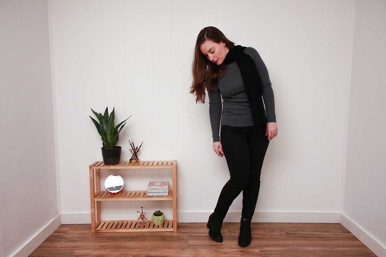 Alyssa wears a grey sweater, black skinny jeans, black booties, and a black scarf while looking down at the floor