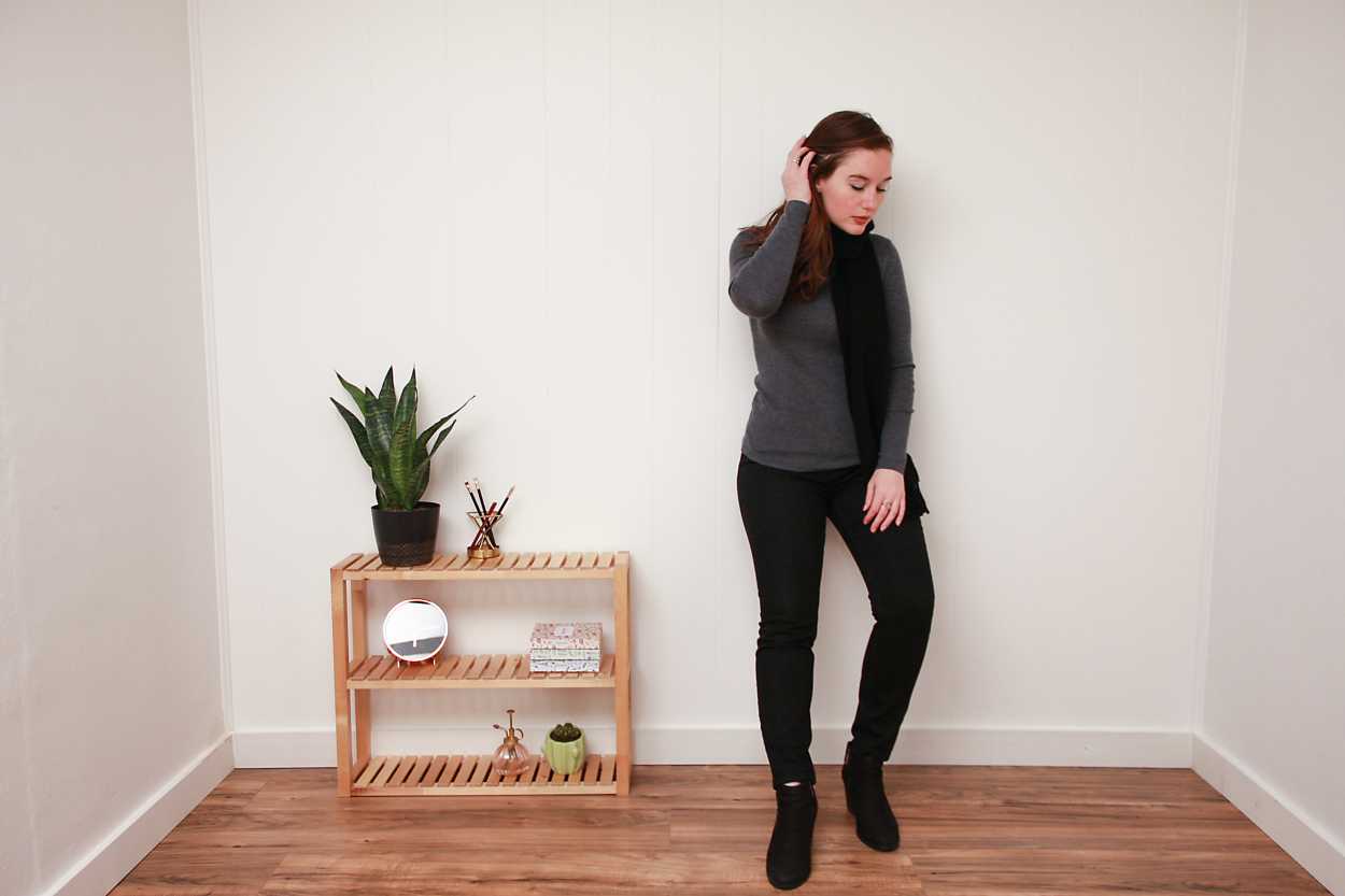 Alyssa wears a grey sweater, black skinny jeans, black booties, and a black scarf while leaning against a wall