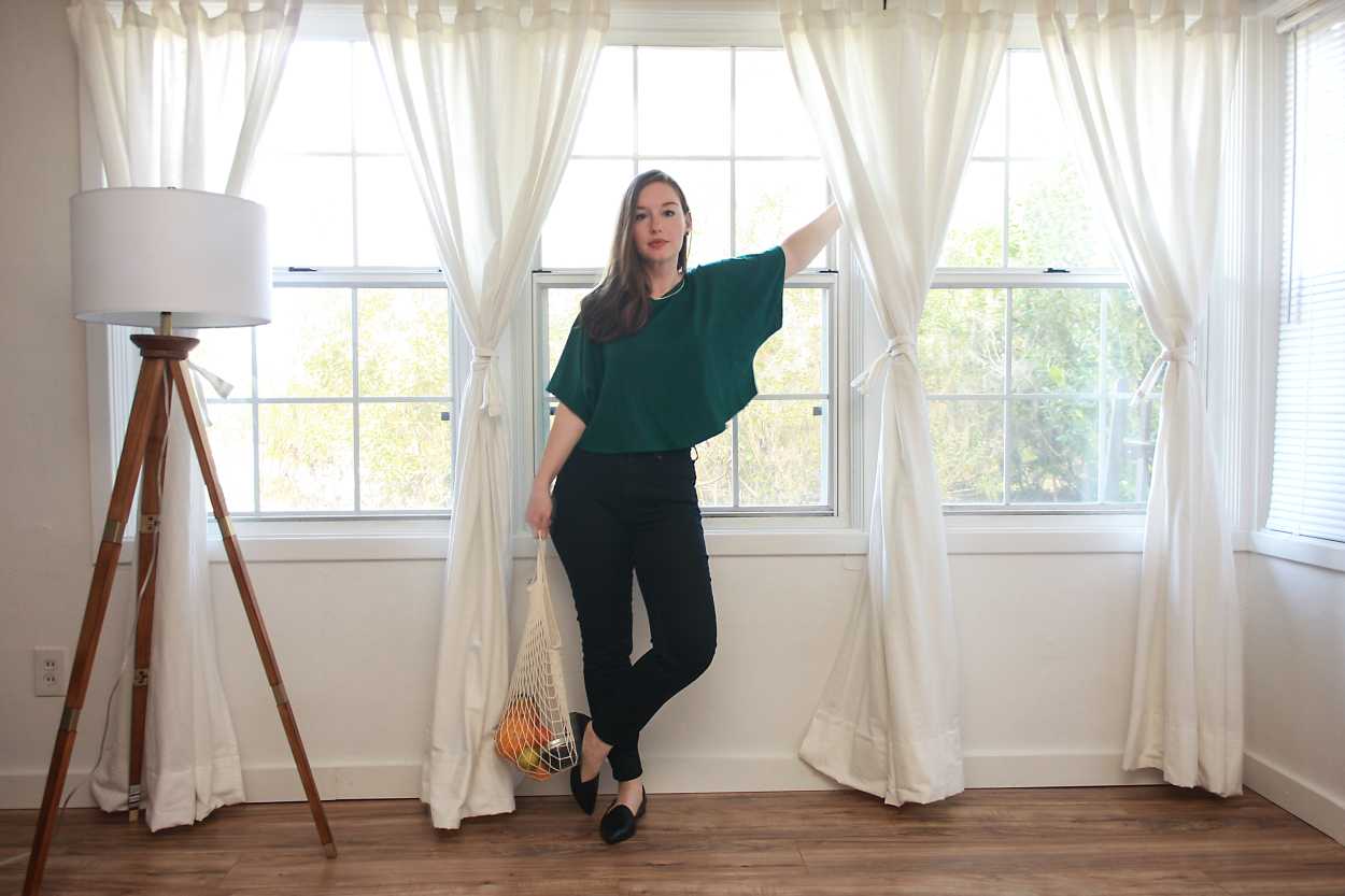 Alyssa wears a green top, black jeans, and black flats while carrying a bag of fruit