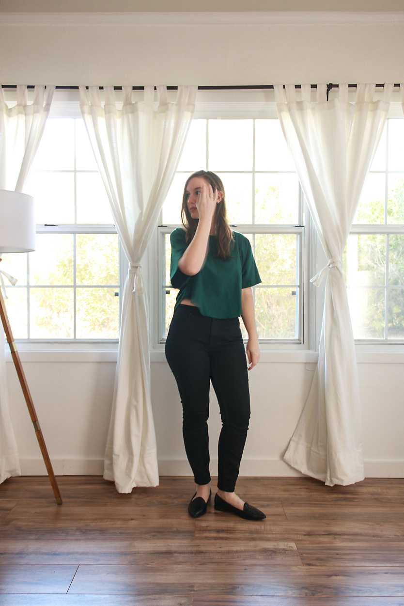 Alyssa wears a green top, black jeans, and black flats while brushing her hair from her face