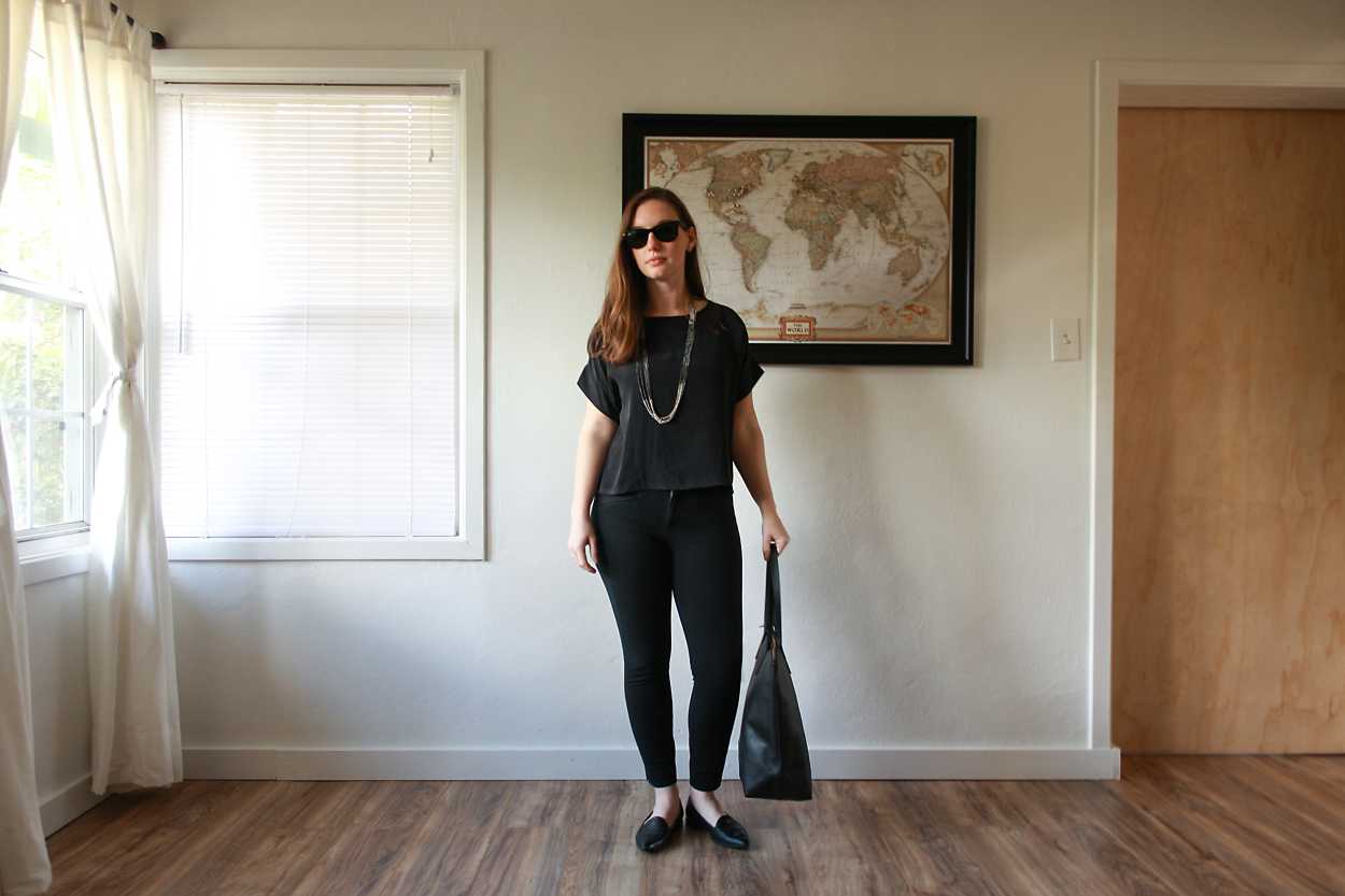 Alyssa wears a silk tee, black pants, and black flats while holding a tote