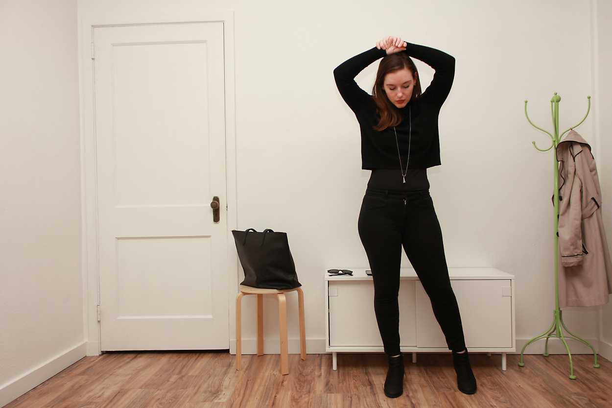 Alyssa wears a black cashmere mockneck sweater with black pants and boots and places her hands on her head