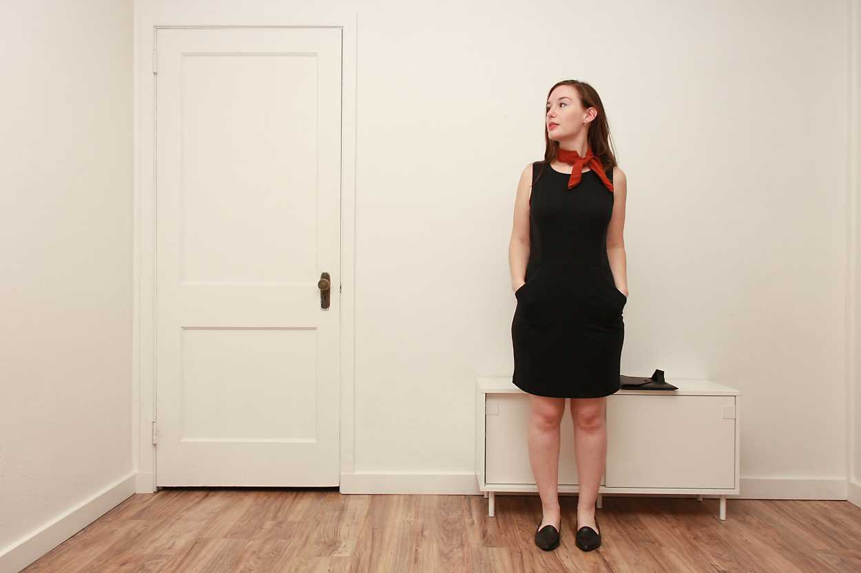 Alyssa wears a black sheath dress with black flats and a rust bandana while placing her hands in her pockets