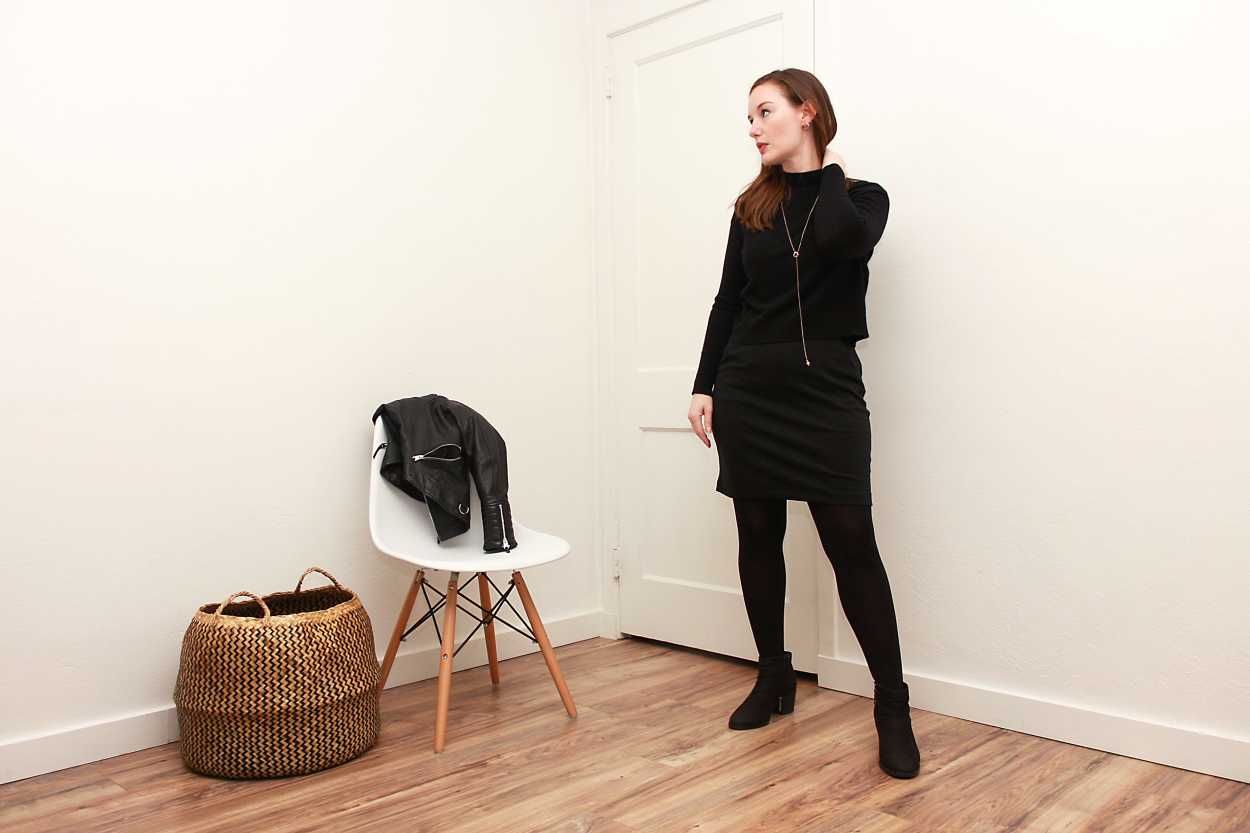Alyssa wears a black turtleneck sweater over a black dress with tights and boots and looks away from the camera