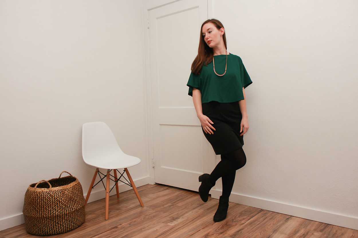 Alyssa wears a green silk top over a black dress with tights and boots and kicks one knee like a flamingo