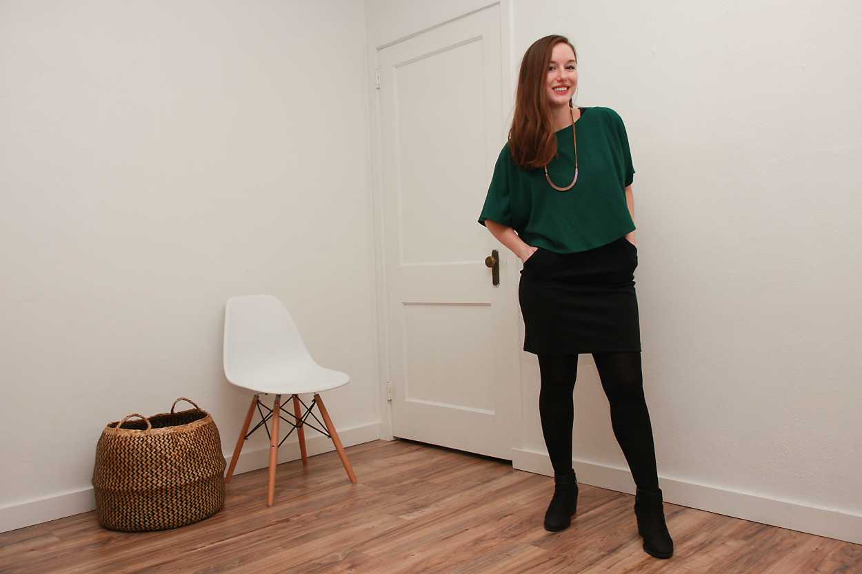 Alyssa wears a green silk top over a black dress with tights and boots and places her hands in her pockets