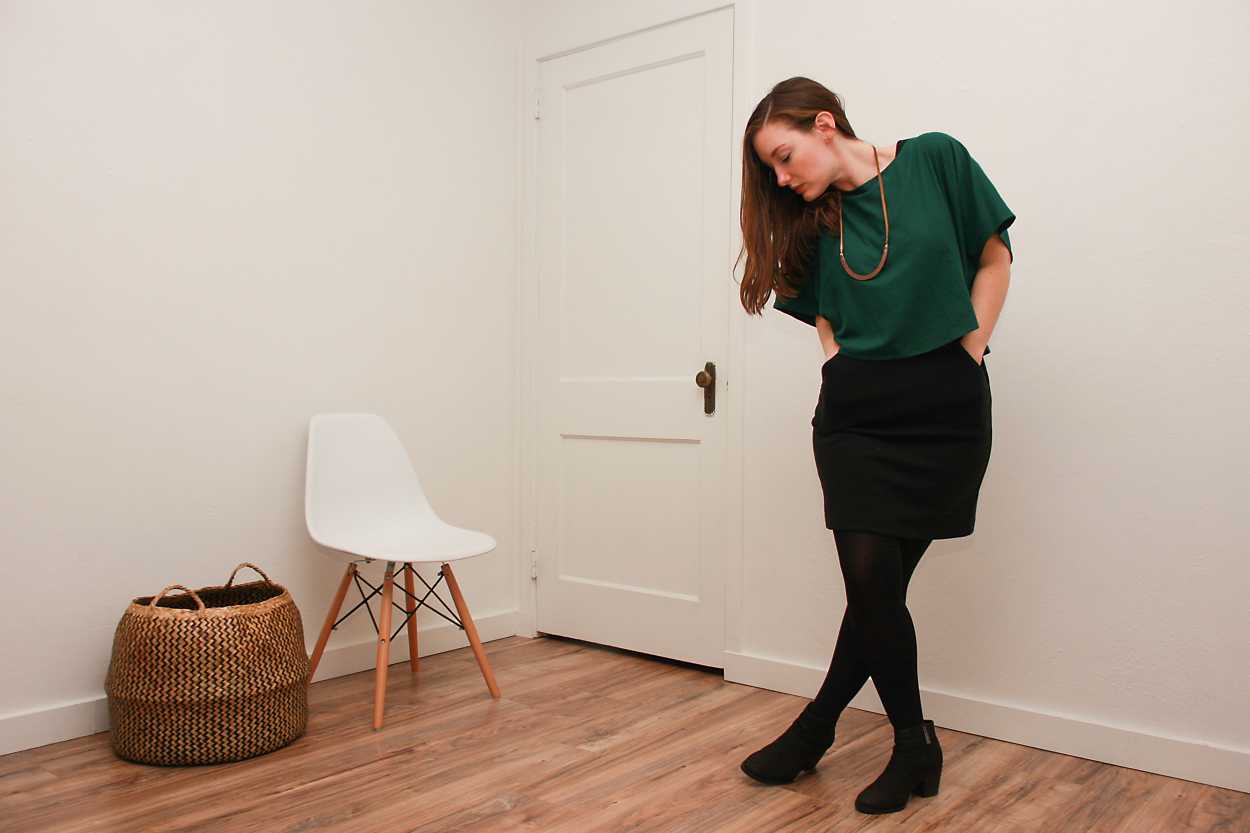 Alyssa wears a green silk top over a black dress with tights and boots while looking downward