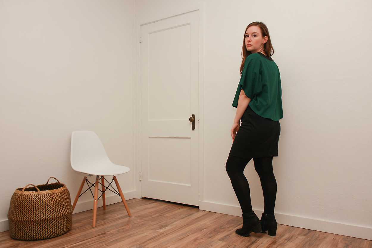 Alyssa wears a green silk top over a black dress with tights and boots and faces away from the camera