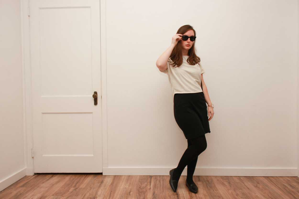 Alyssa wears a white silk tee over a black dress with tights and flats, while placing a pair of sunglasses on her face