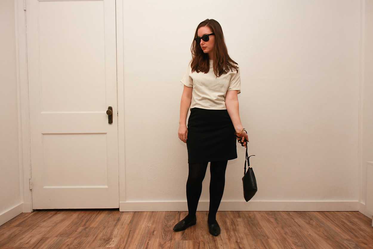 Alyssa wears a white silk tee over a black dress with tights and flats while dangling a purse at her side