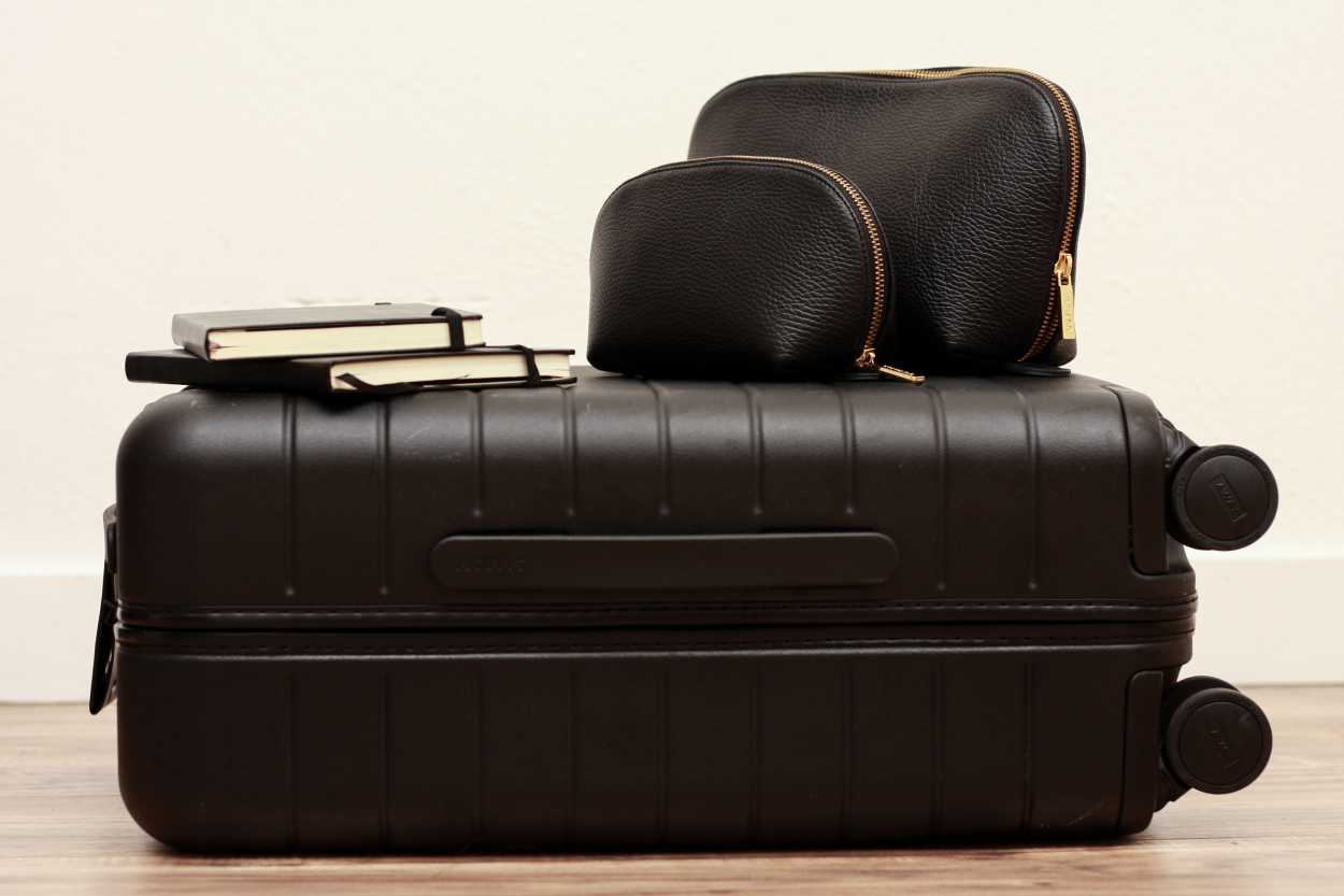 The Leather Travel Case Set from Cuyana sits on top of an Away Carry-On Suitcase with a set of journals
