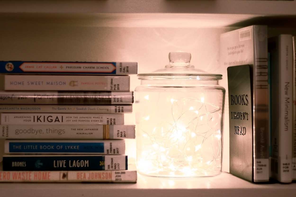 A stack of library books on a shelf next to a jar filled with string lights