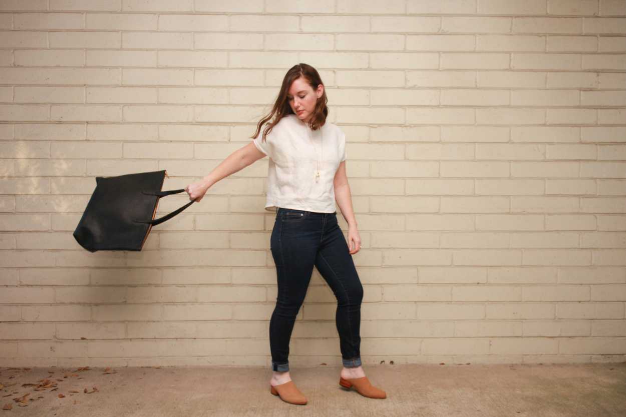 Alyssa wears a homemade white linen tee, skinny blue jeans, and nude mules and swings a tote behind her