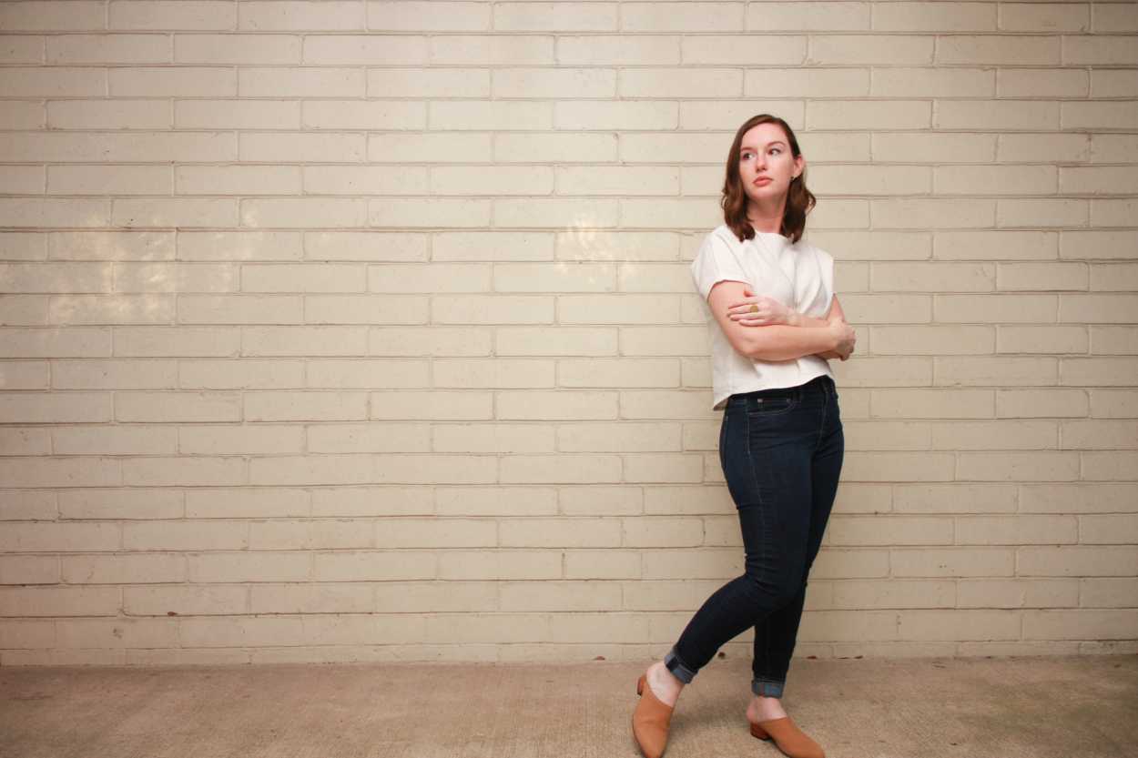 Alyssa wears a homemade white linen tee, skinny blue jeans, and nude mules and stands with her arms folded