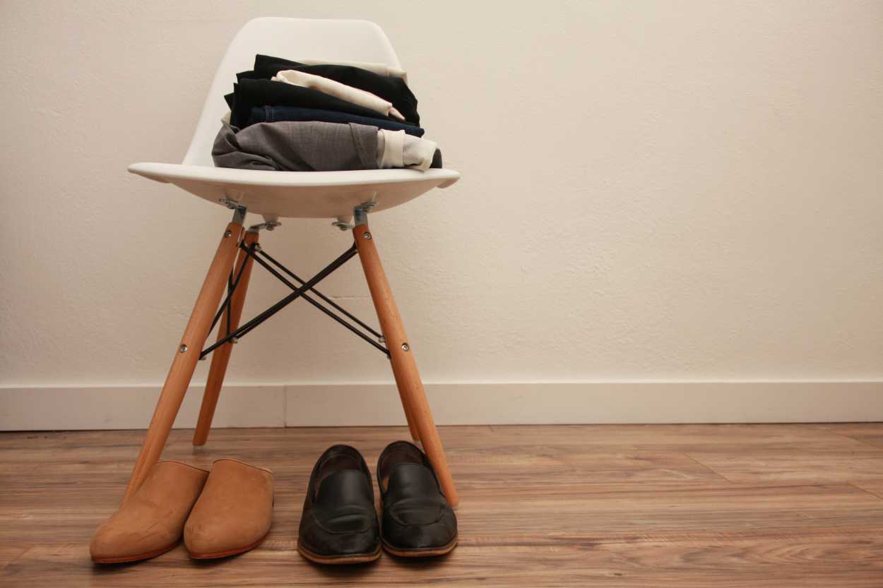 Eight garments and two pairs of shoes sit on and next to a white Eames chair