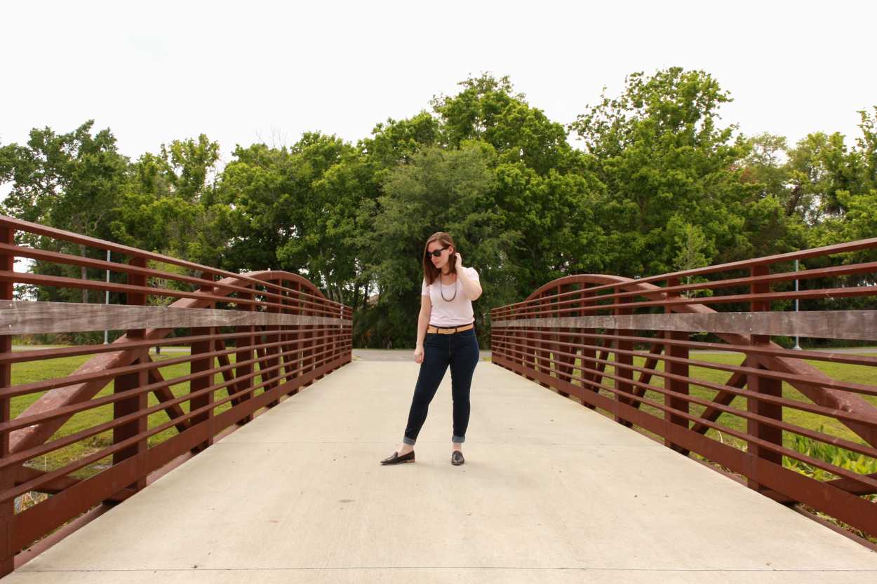 Alyssa wears a pink tee, blue jeans, black loafers, and a tan belt. She is standing on a bridge, and tucking her hair behind her ear.