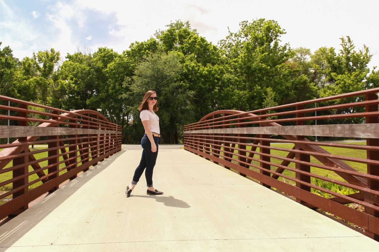 Alyssa wears a pink tee, blue jeans, black loafers, and a tan belt. She is standing on a bridge, facing sideways to the camera.