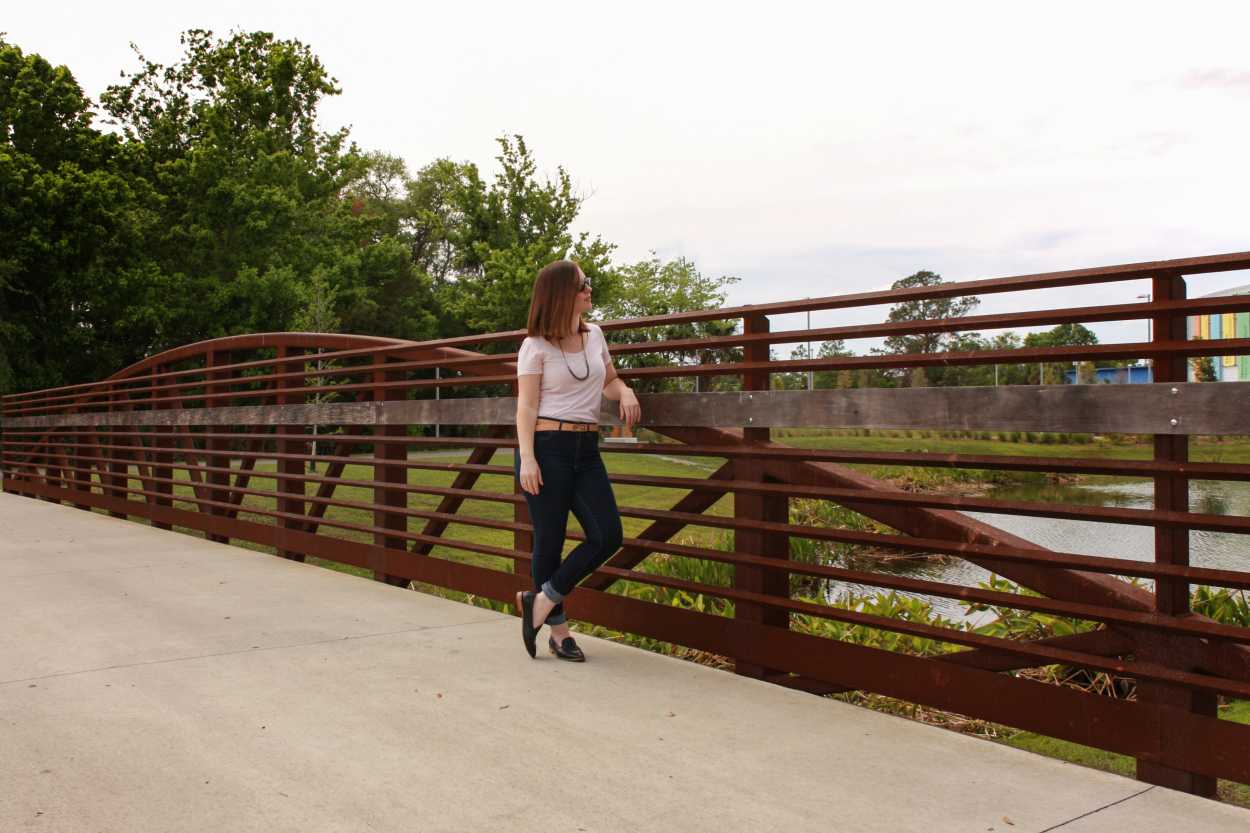 Alyssa wears a pink tee, blue jeans, black loafers, and a tan belt. She is standing on a bridge, and leaning against the railing