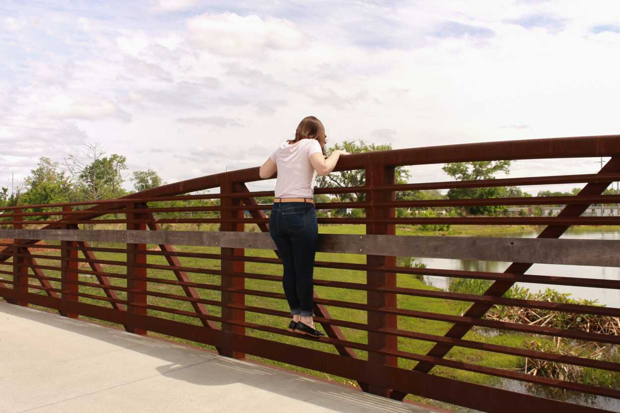 Alyssa wears a pink tee, blue jeans, black loafers, and a tan belt. She is standing on a bridge, and peering over the railing