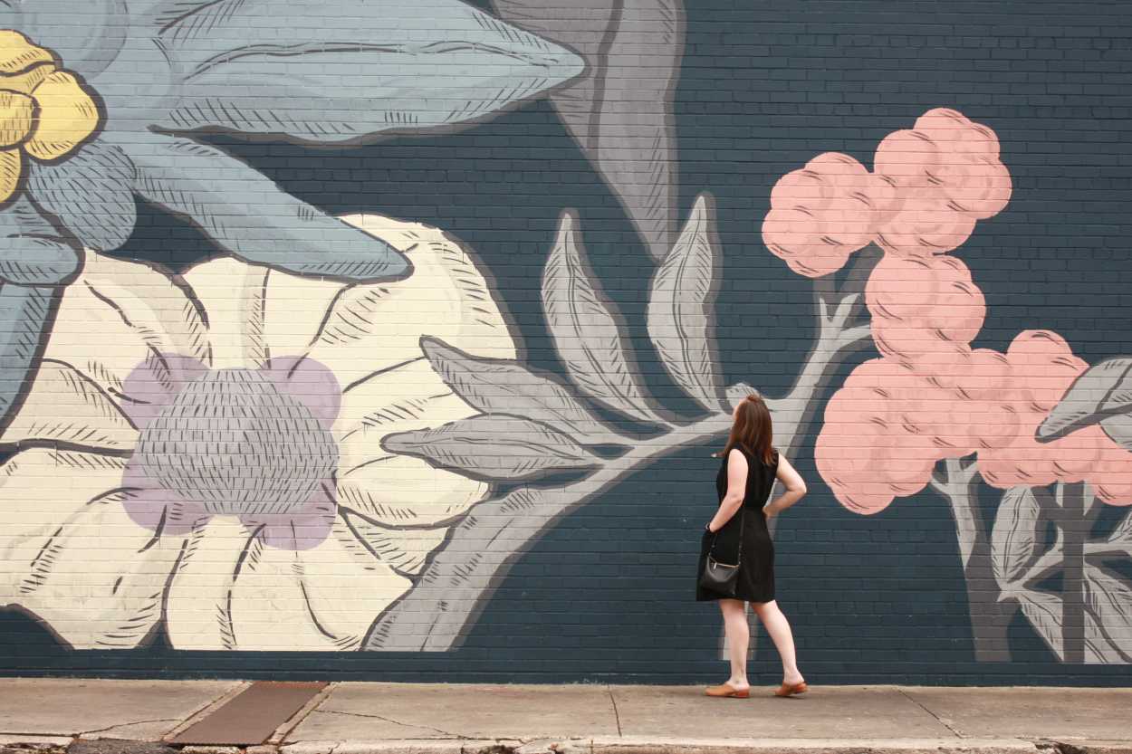 Alyssa wears a white tunic underneath a black dress with tan mules. She is standing in front of a floral wall and facing away from the camera