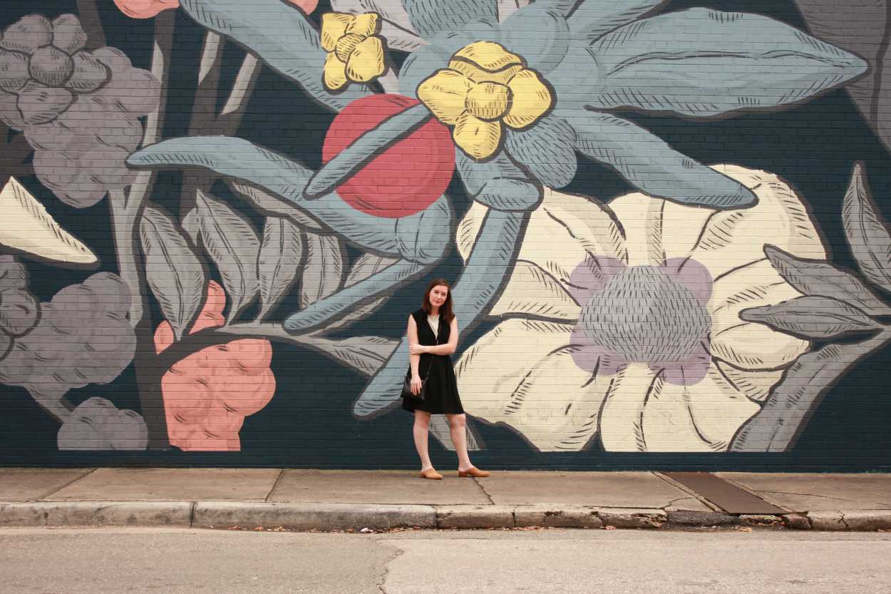 Alyssa wears a white tunic underneath a black dress with tan mules. She is standing in front of a floral wall and holding her arms half crossed