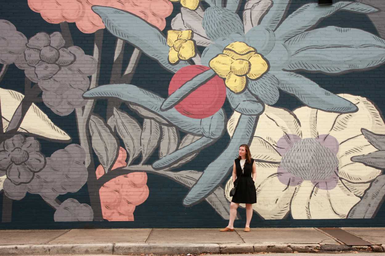 Alyssa wears a white tunic underneath a black dress with tan mules. She is standing in front of a floral wall and looking off to the side