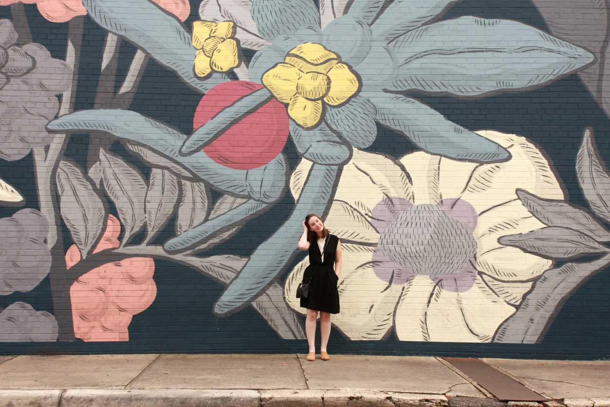 Alyssa wears a white tunic underneath a black dress with tan mules. She is standing in front of a floral wall and running her hand through her hair.