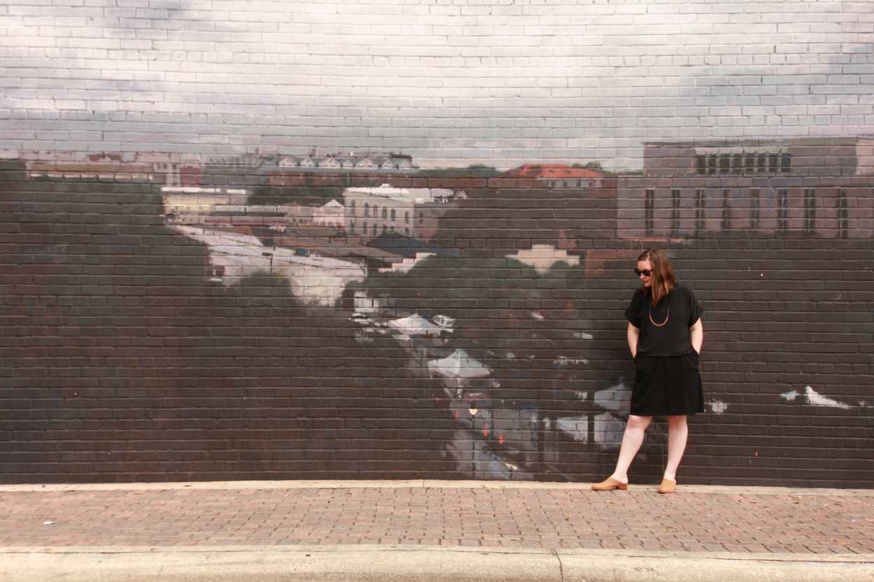 Alyssa wears a silk tee over a dress with tan mules. She is standing in front of a mural of a city, and placing her hands in her pockets.