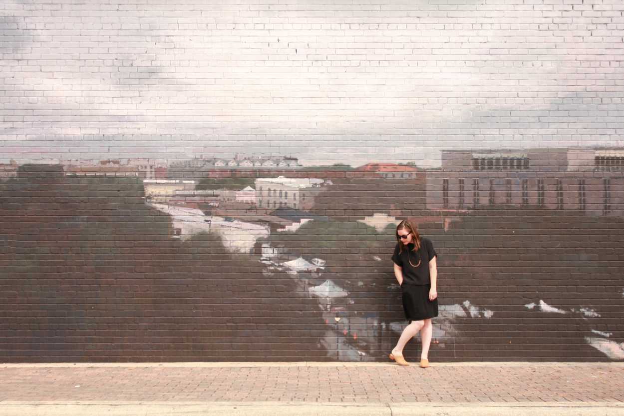 Alyssa wears a silk tee over a dress with tan mules. She is standing in front of a mural of a city, and looking downward.