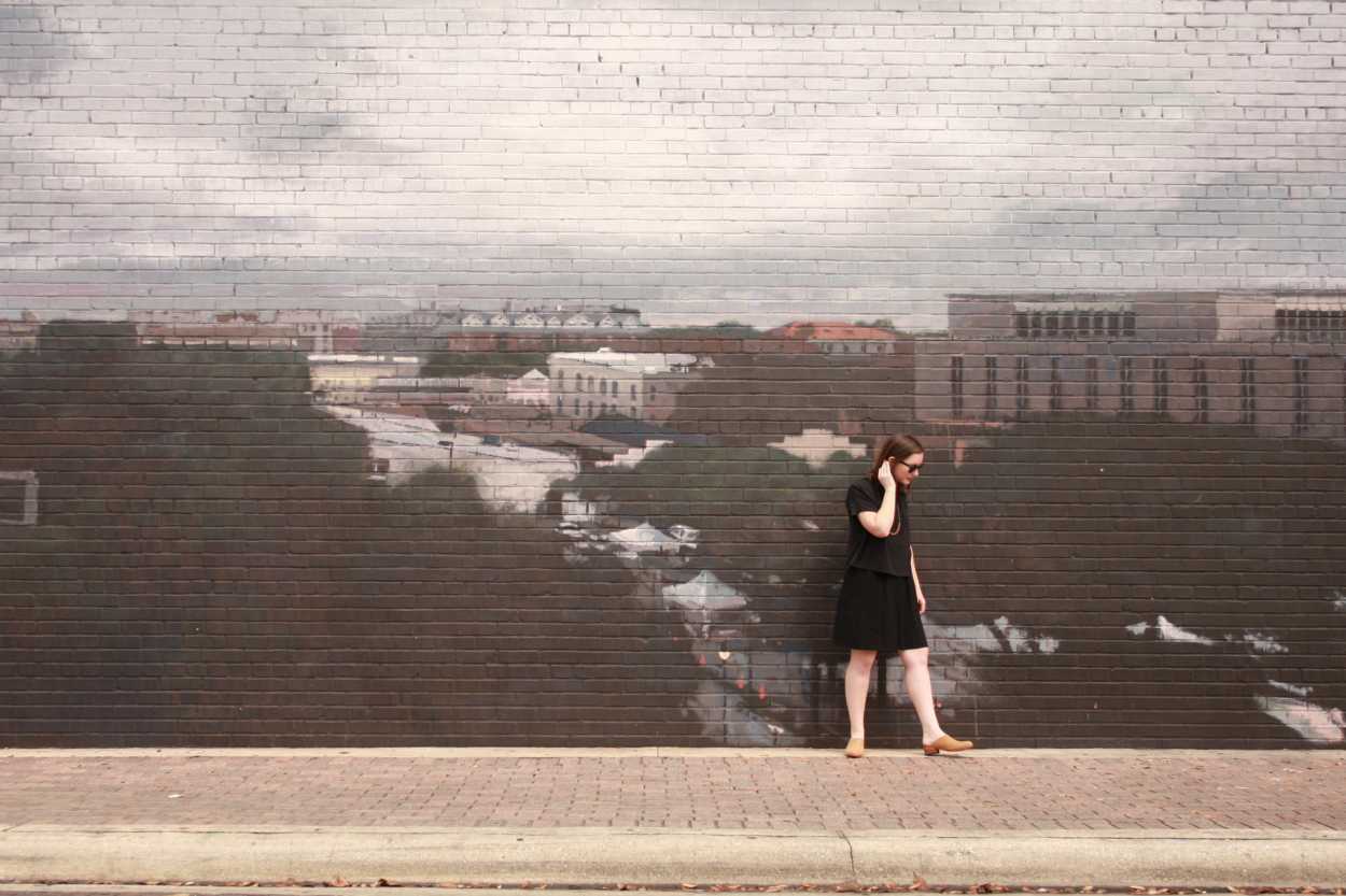 Alyssa wears a silk tee over a dress with tan mules. She is standing in front of a mural of a city, and looking downward while brushing her hair behind her ear.