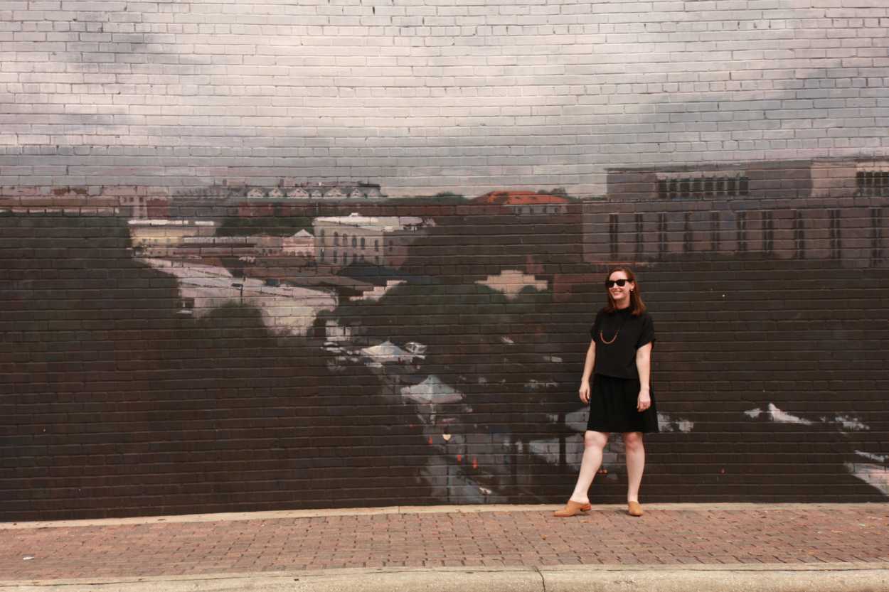 Alyssa wears a silk tee over a dress with tan mules. She is standing in front of a mural of a city, and smiling at the camera.