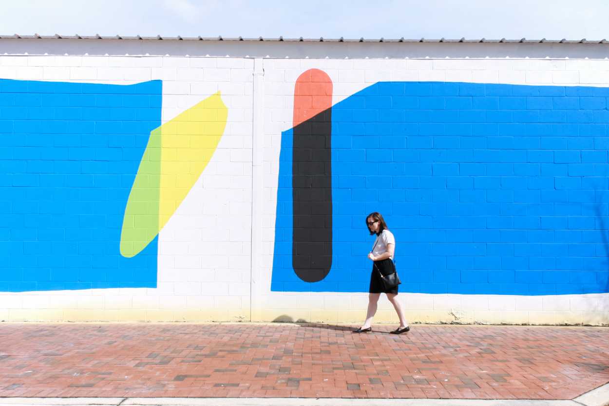Alyssa wears a black dress with a white tee on top, and a pair of black loafers. She walks in front of a colorful mural