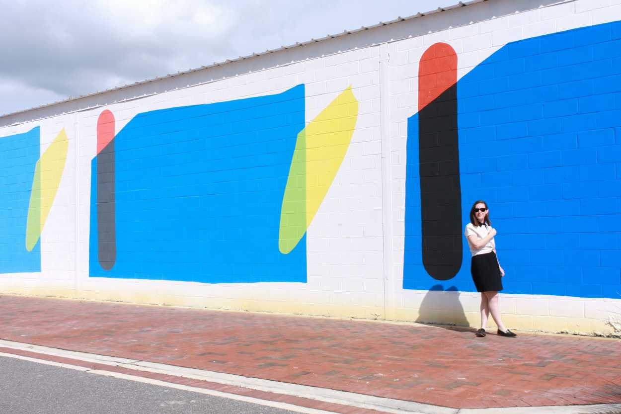 Alyssa wears a black dress with a white tee on top, and a pair of black loafers. She walks in front of a colorful mural, and she is crossing her arm in front of her