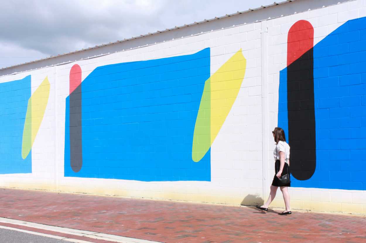 Alyssa wears a black dress with a white tee on top, and a pair of black loafers. She walks in front of a colorful mural, and she is facing away from the camera