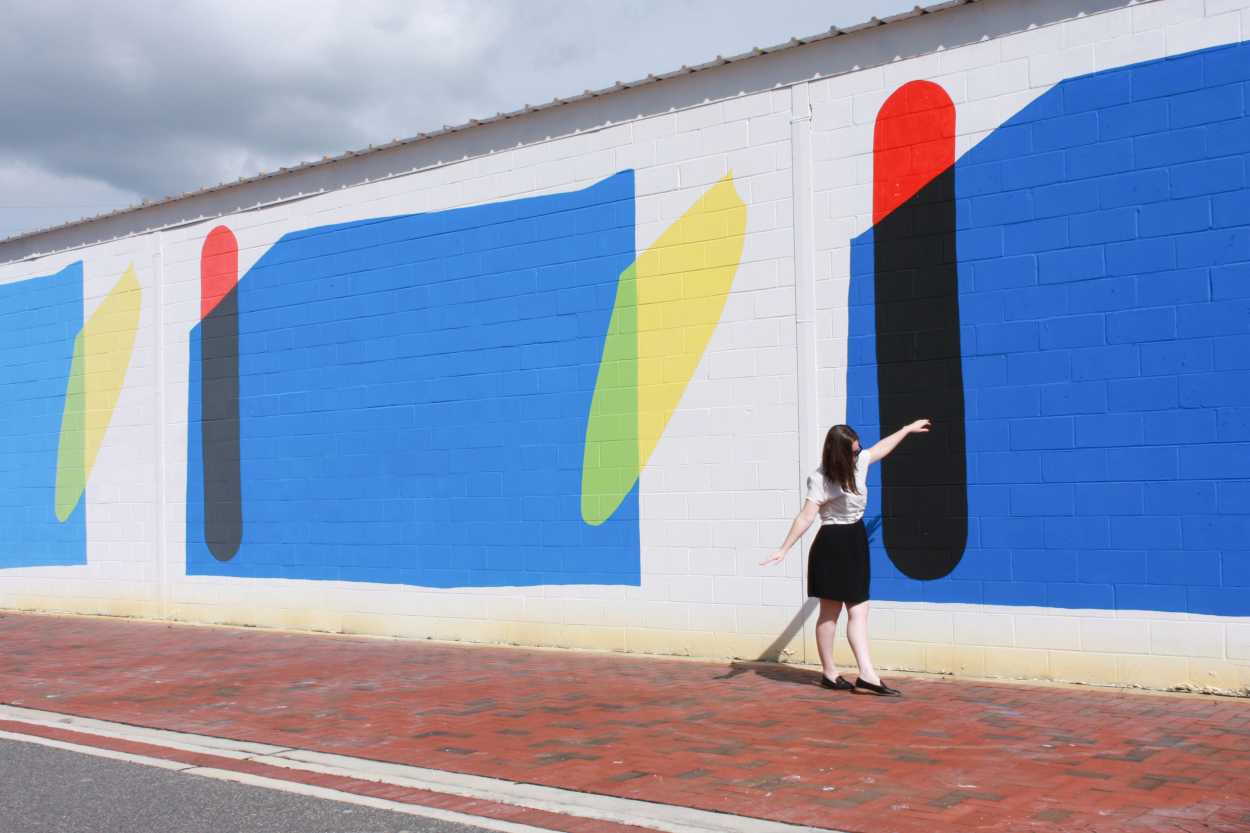 Alyssa wears a black dress with a white tee on top, and a pair of black loafers. She walks in front of a colorful mural, and she is pretending to walk a straight line