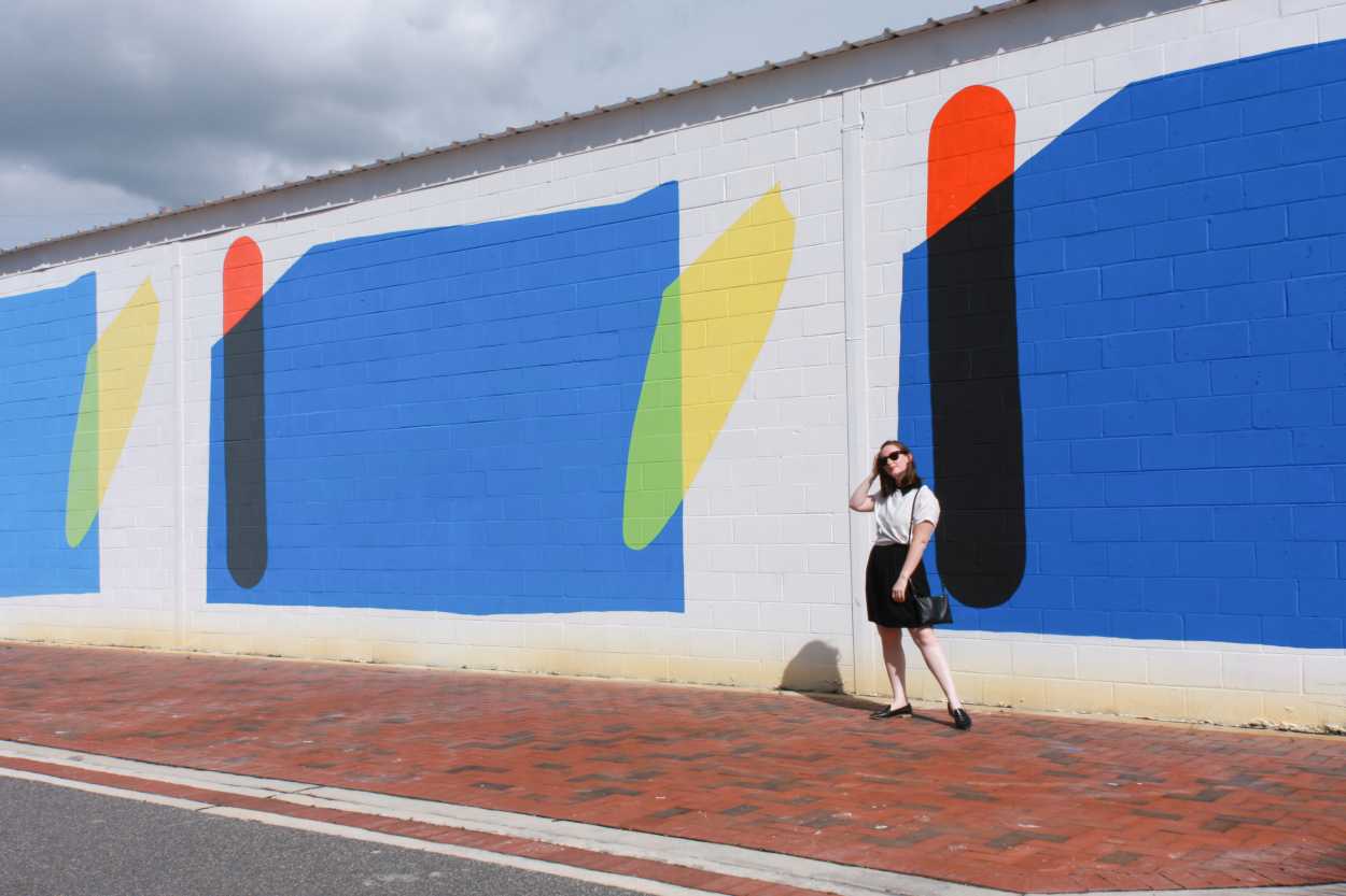 Alyssa wears a black dress with a white tee on top, and a pair of black loafers. She walks in front of a colorful mural, and she is brushing her hair behind her ear