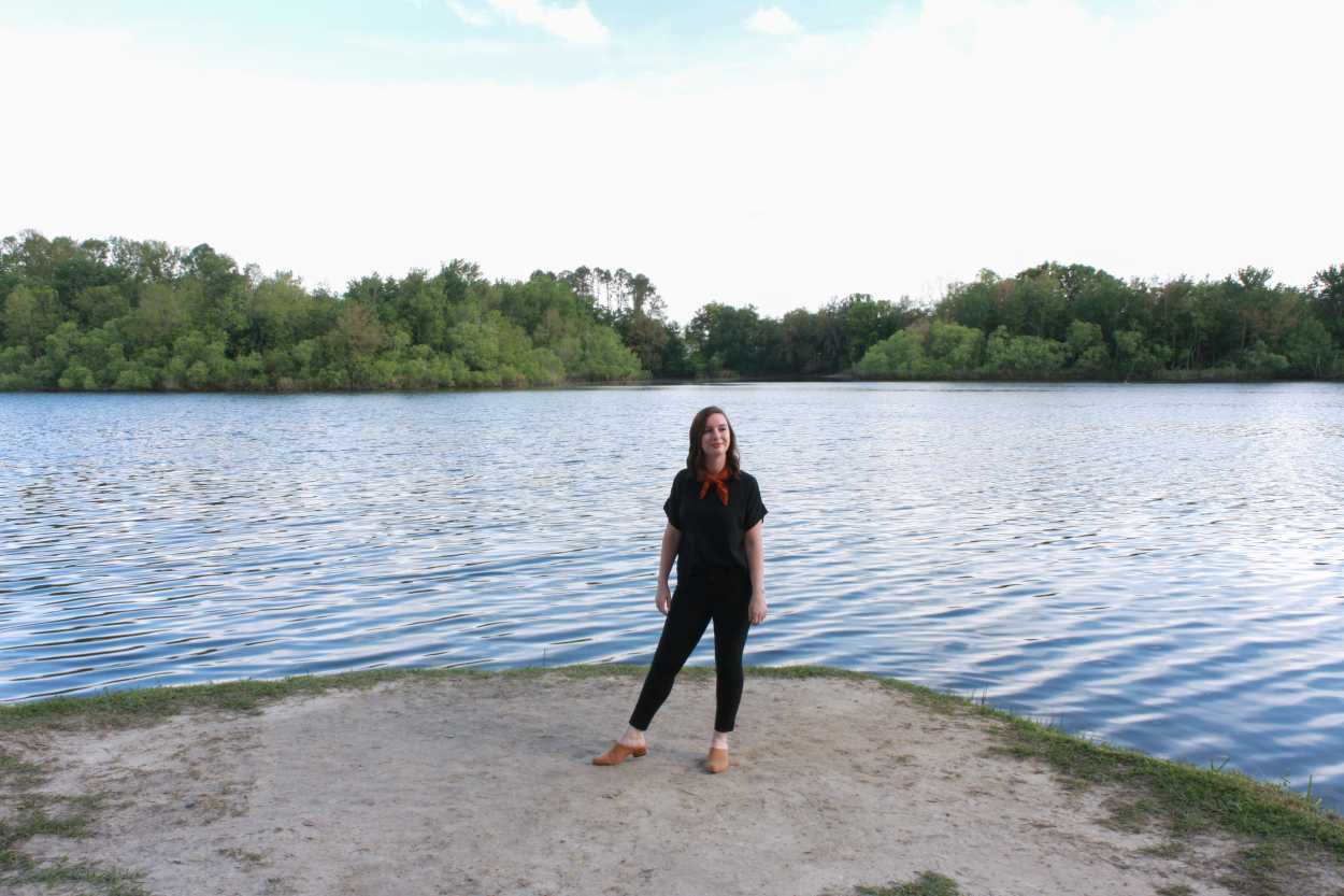 Alyssa is wearing a black silk tee, black pants, tan mules, and a rust bandana. She is standing in front of a lake and facing the camera.