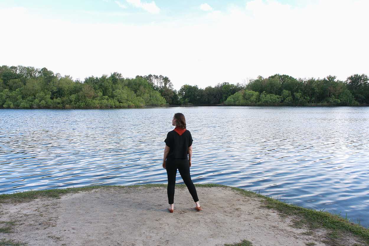 Alyssa is wearing a black silk tee, black pants, tan mules, and a rust bandana. She is standing in front of a lake and facing away from the camera.