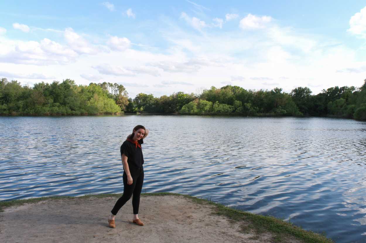 Alyssa is wearing a black silk tee, black pants, tan mules, and a rust bandana. She is standing in front of a lake and running her hand through her hair