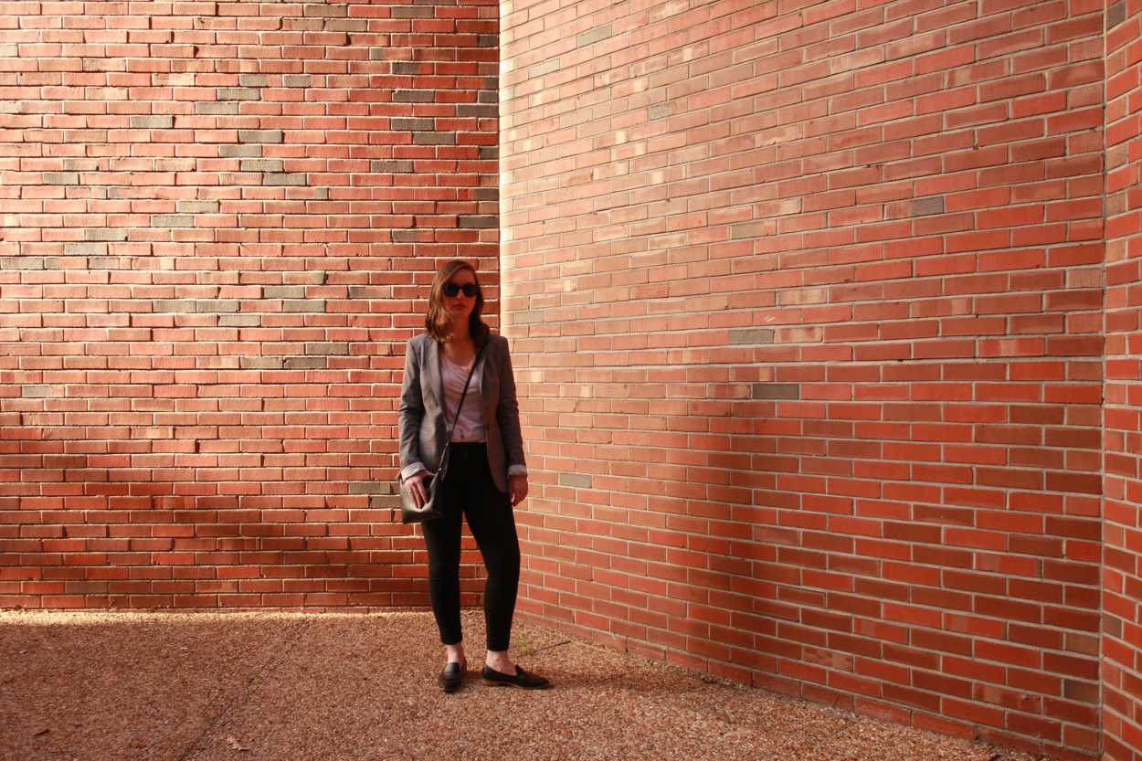 Alyssa wears a blush tee, black pants, black loafers, and a grey blazer. She is by a brick wall, and kind of far from the camera