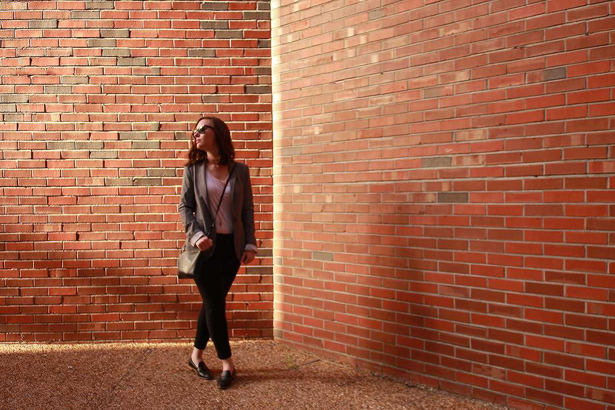 Alyssa wears a blush tee, black pants, black loafers, and a grey blazer. She is by a brick wall, and walking toward the camera.