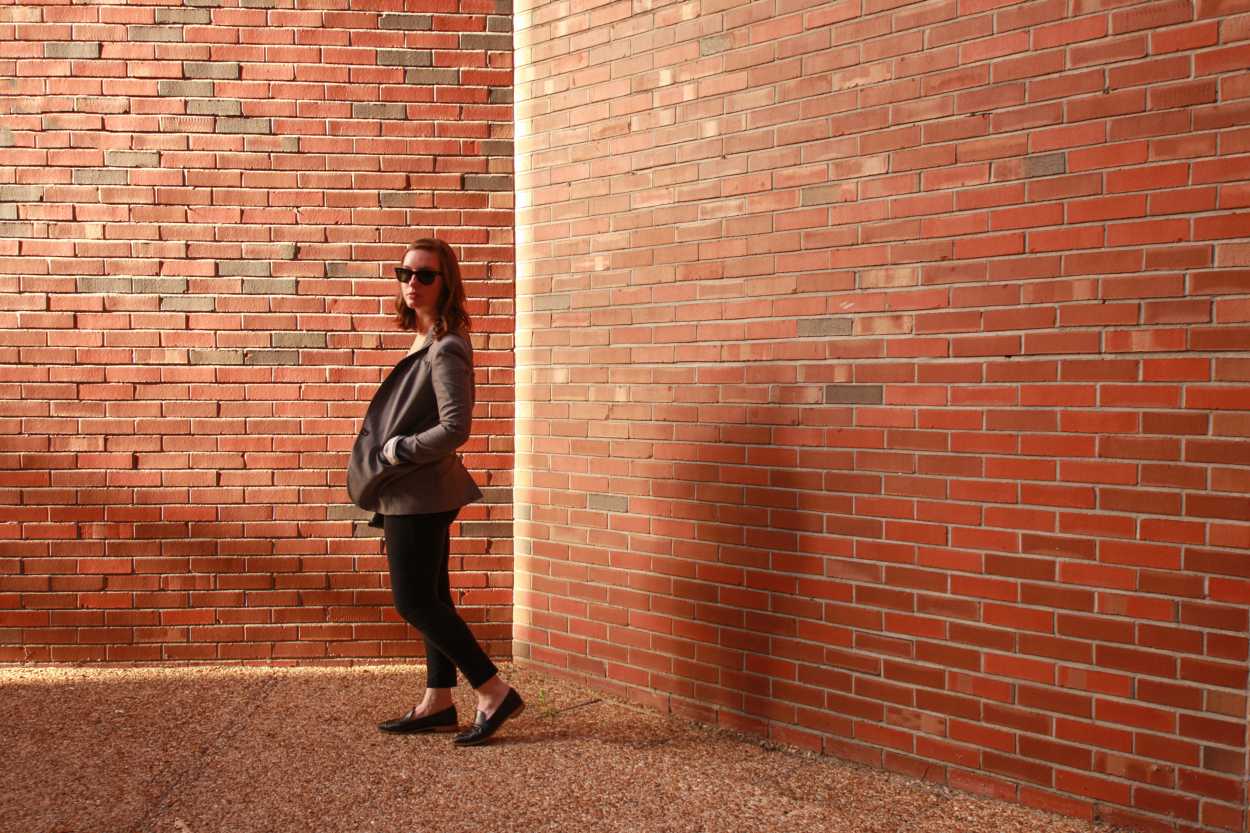 Alyssa wears a blush tee, black pants, black loafers, and a grey blazer. She is by a brick wall, facing sideways to the camera.
